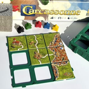 Könnte beinhalten: Das Brettspiel "Carcassonne" mit farbigen Holzspielsteinen, grünen Kartenhaltern und Landschaftskarten mit Straßen, Städten und Feldern. Ein Stapel Karten ist ebenfalls zu sehen.