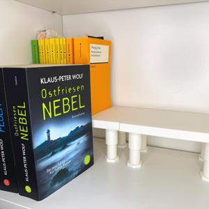 Könnte beinhalten: Bücher auf einem weißen Regal. Die Bücher haben schwarze Einbände mit deutschen Titeln, darunter "Ostfriesen Nebel" von Klaus-Peter Wolf. Weitere Bücher sind in Gelb und Orange sichtbar. Ein weißes Regal mit verstellbaren Beinen ist ebenfalls vorhanden.