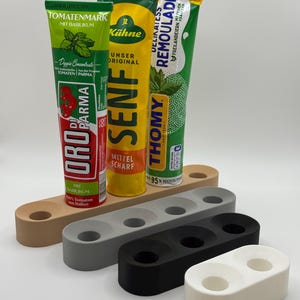Könnte beinhalten: Eine Auswahl an Würzmittel-Tuben, darunter Tomatenmark, Senf und Remoulade, wird auf einer weißen Oberfläche präsentiert. Die Tuben sind auf rechteckigen Haltern in Beige, Grau, Schwarz und Weiß platziert. Die Tuben haben Text auf Deutsch.