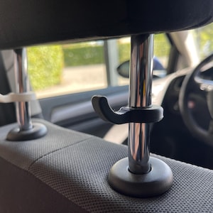 Könnte beinhalten: Kopfstütze im Auto mit schwarzer Kopfstütze und silberner Metallstütze. Zwei Haken, einer weiß und einer schwarz, sind an der Stange befestigt. Die Haken sind zum Halten von Gegenständen im Auto bestimmt.