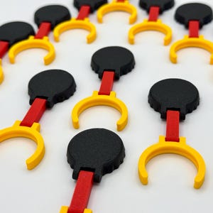 K&ouml;nnte beinhalten: Eine Ansammlung kleiner, mehrteiliger Komponenten, die in einer Gruppe angeordnet sind. Jedes Teil besteht aus einem runden schwarzen Oberteil, einem geraden roten Verbindungssteg und einem gelben C-f&ouml;rmigen Clip an der Basis.