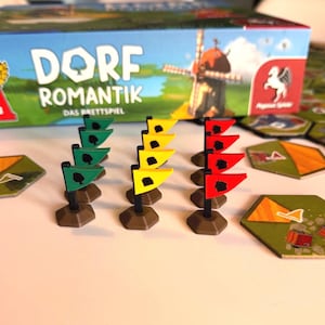 Könnte beinhalten: Das Brettspiel "Dorf Romantik" mit einer Schachtel, die eine Windmühle zeigt. Das Spiel enthält grüne, gelbe und rote Flaggen und sechseckige Spielsteine. Auf der Schachtel steht "Spiel des Jahres 2022".