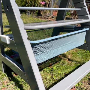 Könnte beinhalten: Ein grauer Hochstuhl aus Holz mit einem hellblauen Netzkorb und einem transparenten Kunststofftablett. Der Hochstuhl steht im Freien auf einer Rasenfläche.
