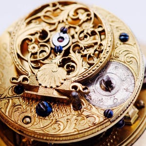 Könnte beinhalten: Nahaufnahme eines antiken goldenen Taschenuhrwerks. Das aufwendige Design zeigt kunstvolle Filigranarbeiten, kleine blaue Juwelen und ein silberfarbenes Zifferblatt. Die Uhr ist sehr detailliert und zeigt die Handwerkskunst der Zeit.