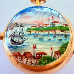 May include: A gold-toned pocket watch with a hand-painted seascape scene. The watch face depicts a sailing ship, buildings, and boats on a body of water, under a blue and orange sky. The watch has a gold-colored loop at the top.