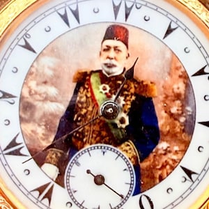 May include: Antique gold pocket watch with a white face and black Roman numerals. The watch face features a portrait of a man in formal attire. The watch has a small seconds dial and ornate gold detailing around the bezel.