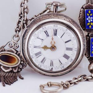 May include: Vintage silver pocket watch with a white face, black Roman numerals, and intricate hands. The watch has an elaborate chain and fob. The chain features blue and yellow enamel details. A classic timepiece.