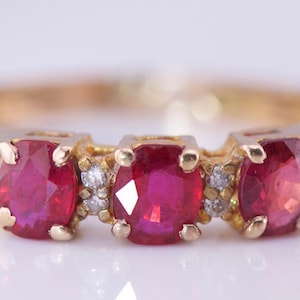 Pode incluir: Um anel de ouro com três pedras vermelhas vibrantes de corte oval, provavelmente rubis, dispostas em linha. Pequenos diamantes são intercalados entre os rubis. O anel tem um design clássico e elegante.