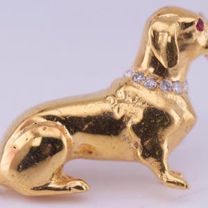 Pode incluir: Estatueta de cão dachshund dourado com uma coleira de pedras preciosas transparentes. O cão tem olhos de pedras preciosas vermelhas e está sentado. A estatueta é provavelmente um broche ou pingente.
