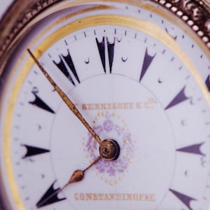 May include: Close-up of an antique pocket watch with a white face, gold hands, and black numerals. The watch face features the text "SERKEKOFF K.C." and "CONSTANTINOPLE" in gold lettering, with a decorative purple floral design.