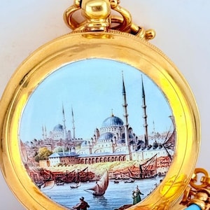 May include: A gold pocket watch with a detailed painted scene of a city with minarets, boats, and figures. The watch has a gold chain and a decorative top. The watch face features a blue sky and a detailed cityscape.