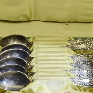 May include: A set of silver spoons in a green satin-lined box. The spoons have ornate handles and are arranged neatly. The box is black with a green trim, suggesting a vintage or antique set, perfect for serving.