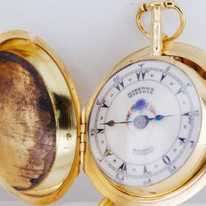 May include: A gold pocket watch with an open lid, revealing a white face with black numerals and hands. The watch face has the text "MARKWICK MARKHAM LONDON". A small red jewel is on the side.