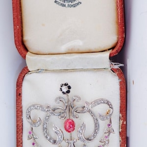 Op de afbeelding: Een antieke broche in een rode en witte presentatiedoos. De broche heeft een centrale rode edelsteen, omringd door heldere en roze edelstenen, en sierlijk zilverkleurig metaalwerk. De doos heeft de tekst "FABERGE" erop.