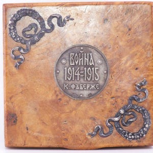 May include: A square, wooden box with a light brown, textured surface. The box features a circular, silver-colored emblem in the center with the text "Война 1914-1915 К. Фаберже". Decorative silver-colored scrollwork adorns the corners.