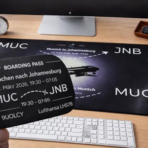 Könnte beinhalten: Ein schwarzer Bordkarten-Schlüsselanhänger mit Flugdetails von München nach Johannesburg. Eine schwarze Mausunterlage mit denselben Informationen und einer Flugzeuggrafik ist ebenfalls zu sehen. Die Artikel liegen auf einem Holzschreibtisch mit Tastatur und Kopfhörern.