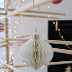 Może przedstawiać: Biała papierowa ozdoba choinkowa w kształcie kuli z wzorem plastra miodu. Ozdoba jest zawieszona na sznurku na drewnianej choince.