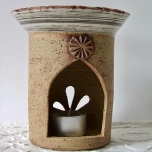 Könnte beinhalten: Ein beigefarbener Keramik-Diffusor für ätherische Öle mit einem dekorativen Blumendesign und einer bogenförmigen Öffnung. Die Oberseite hat ein gestreiftes Muster. Eine brennende Teelichtkerze ist im Inneren sichtbar.