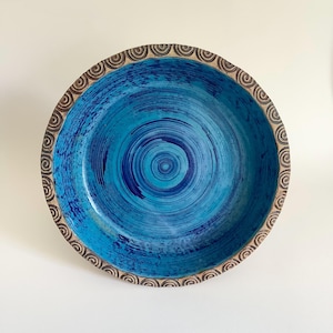 Könnte beinhalten: Eine runde Keramikschale mit einem leuchtend blauen Innenraum und sichtbaren kreisförmigen Pinselstrichmustern. Der breite, flache Rand ist in einem natürlichen Beigeton gehalten und mit einem wiederkehrenden Spiralmuster verziert.