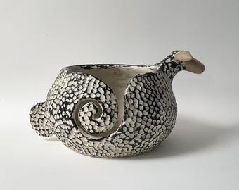 A wool pot, also called a yarn bowl, in the shape of a sheep. The yarn pot is made of stoneware clay and is black and white.