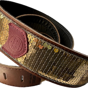 May include: A guitar strap featuring a wide band covered in shimmering gold sequins. The strap includes a dark red leather patch embossed with the text Copper Pence, and a brown leather end piece with pre-cut slots for attachment to a guitar.