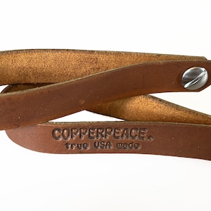 May include: Two brown leather straps with silver metal rivets. The straps are folded in half and the text "COPPERPEACE. true USA made" is embossed on one strap.