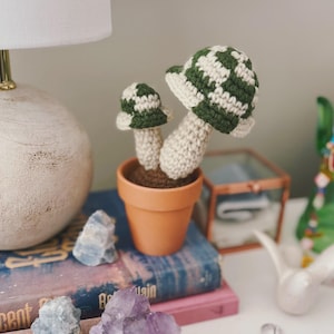 May include: A small terracotta pot holds two crocheted mushrooms. The mushroom caps feature a green and white checkered pattern. The stems are off-white, and the pot is filled with brown soil. The mushrooms are a whimsical decorative item.