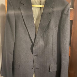 May include: A charcoal gray blazer with a two-button closure, notched lapels, and two front pockets. The blazer is hanging on a wooden hanger against a dark wood door. The interior lining is a light tan color.