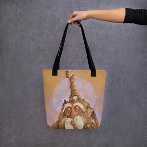 Könnte beinhalten: Tragetasche mit einer detaillierten Illustration eines alten Charakters mit langem weißem Bart und hohem, strukturiertem Hut. Das Kunstwerk ist vor einem weichen, kreisförmigen Hintergrund mit hellbraunem und violettem Farbschema platziert. Die Tasche hat schwarze Griffe.