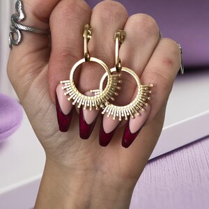 Puede incluir: Pendientes de aro dorados con un diseño de sol. Los pendientes se muestran en una mano con esmalte de uñas rojo y rosa.