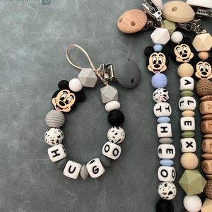 May include: A collection of four pacifier clips featuring silicone and wooden beads in shades of blue, gray, white, and tan. The clips include character beads resembling Mickey Mouse, and white letter beads spelling out the names HUGO, THEO, ALEX, and MAXIM.