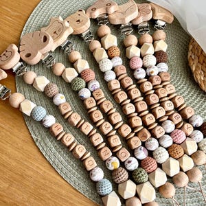 Op de afbeelding: Houten fopspeenclips voor baby's met dierfiguren, zoals een beer en een olifant. De clips hebben houten kralen, gehaakte kralen en blokken met letters met namen. Op een groene placemat. Namen: Hugo, Romi, etc.
