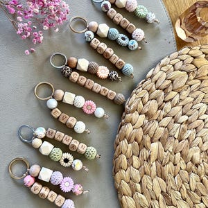 Könnte beinhalten: Eine Sammlung personalisierter Schlüsselanhänger mit Holzperlen und Buchstabenblöcken, die Namen wie "Kate" und "Max" buchstabieren. Jeder Schlüsselanhänger hat dekorative Elemente wie Blumen und gemusterte Perlen. Die Schlüsselanhänger sind auf einer neutralen Oberfläche angeordnet.