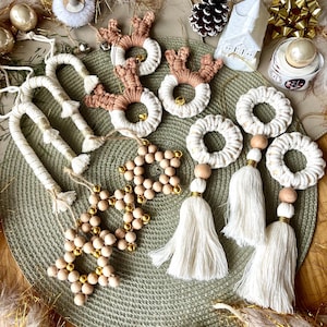 Op de afbeelding: Een verzameling handgemaakte kerstversieringen met verschillende ontwerpen, waaronder rendieren, sterren en cirkels. De versieringen zijn gemaakt van natuurlijke materialen zoals houten kralen en macramé, en zijn versierd met gouden accenten.