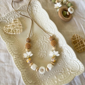 Peut inclure: Un collier composé de perles fleurs en silicone blanc, de sphères crochetées beige, de formes géométriques en bois lisse et de perles à motifs floraux sur un cordon fin. Deux cœurs en rotin tressé reposent sur un plateau en céramique gaufrée.