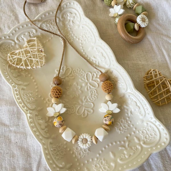 May include: A necklace featuring a mix of white silicone flower beads, tan crocheted spheres, smooth wooden geometric shapes, and floral patterned beads on a thin cord. Two woven rattan hearts are placed on a white embossed ceramic tray.