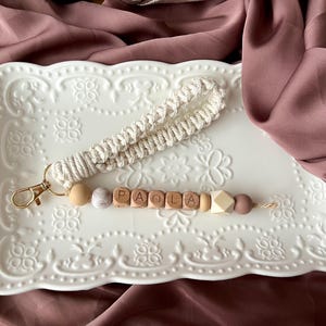 Peut inclure: Un cordon de poignet fait à la main avec le nom "PAOLA" épelé en perles de bois. Le cordon comprend une sangle de poignet en macramé crème, un fermoir doré et des perles décoratives aux tons neutres. Le cordon repose sur un plateau blanc décoratif.