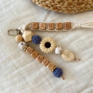 Può includere: Due portachiavi fatti a mano e un accessorio con perline sono esposti su una superficie di lino neutra. Un portachiavi recita "PAULO" con perline di lettere di legno. L'altro portachiavi recita "MAMIE". Gli accessori includono perline di legno, un fiore e una foglia.