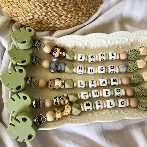 Può includere: Cinque clip per ciuccio con clip in silicone verde salvia a forma di foglia, chiusure in metallo e perline a forma di animale. Ogni clip include perline bianche con lettere che compongono i nomi JADE, HUGO, LIAM, THEO e MILO, arricchite da perline in silicone rotonde e a forma di foglia in tonalità neutre e verdi.