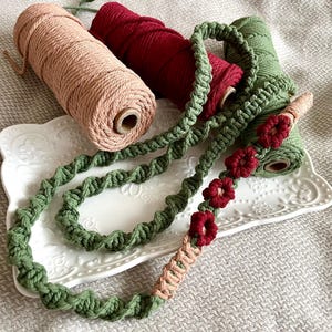 Op de afbeelding: Een groene macramé koord met rode bloemenaccenten wordt getoond op een wit bord met drie spoelen koord in roze, rood en groen.