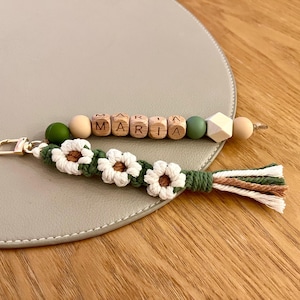 Peut inclure: Un porte-clés fait à la main avec le nom "MARIA" épelé en perles de lettres en bois. Le porte-clés présente un design macramé avec des accents de fleurs blanches et une gland. La palette de couleurs comprend le vert, le blanc et le beige.