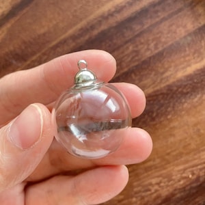 May include: A small, spherical, clear glass ornament featuring a smooth, transparent surface and a polished silver-toned metal cap at the top with a small circular loop for hanging. The object is hollow and round, designed as a decorative pendant.