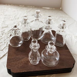 May include: A selection of seven clear glass perfume bottles with decorative stoppers, displayed on a dark brown wooden tray. The bottles have various shapes and sizes, including rounded and angular designs.