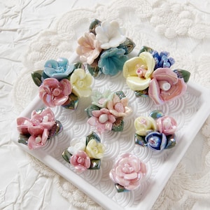 May include: A collection of colorful ceramic flower brooches in various pastel shades, including pink, blue, yellow, and white. Each brooch features detailed petal designs and green leaf accents, arranged on a white tray.