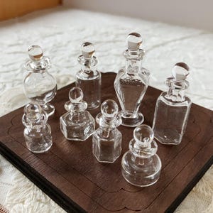 Puede incluir: Una colección de ocho botellas de vidrio transparente en miniatura con tapones decorativos. Las botellas varían en forma, incluyendo diseños redondos, cuadrados y hexagonales. Están dispuestas sobre una bandeja de madera marrón oscuro, mostrando su delicada artesanía.