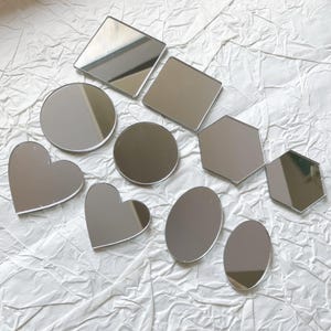 May include: A collection of reflective acrylic shapes, including squares, circles, hearts, ovals, and hexagons. The shapes are silver and arranged on a textured white surface. These decorative pieces are suitable for crafts and DIY projects.