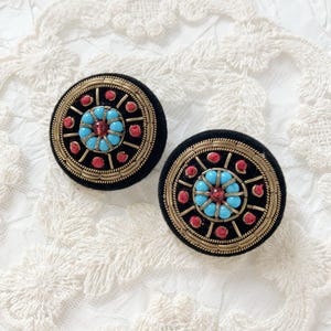 May include: Pair of black velvet round earrings with gold trim and intricate beadwork. The earrings feature a central cluster of turquoise beads surrounded by red and gold accents, creating a striking contrast. The design is reminiscent of a floral or mandala pattern.