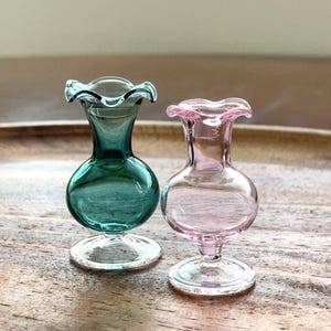 May include: Two miniature glass vases with ruffled rims and pedestal bases. One vase is a deep teal green, and the other is a soft translucent pink. Both items feature a rounded body and a narrow neck, crafted from smooth, clear glass material.