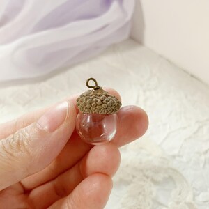 May include: A miniature glass acorn pendant with a brown cap and a small metal loop. The acorn is held in a hand. The pendant is a nature-inspired accessory. The background is soft and blurred.