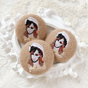 May include: Three round, tan-colored embroidered patches with a portrait of a person wearing a white hat and a tan uniform. The portrait features black hair, red highlights, and a smiling face. The patches are arranged on a white, ornate tray.
